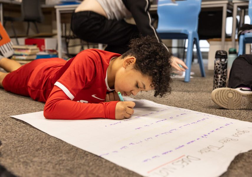 A young boy is lying on the floor in a classroom writing on some big paper with a felt tip